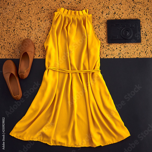 Layout with clothes in brown, orange, yellow warm tones. Feminine beautiful stylish mustard dress with open shoulders. Simple brown shoes without heel and unusual black notebook in form of camera.