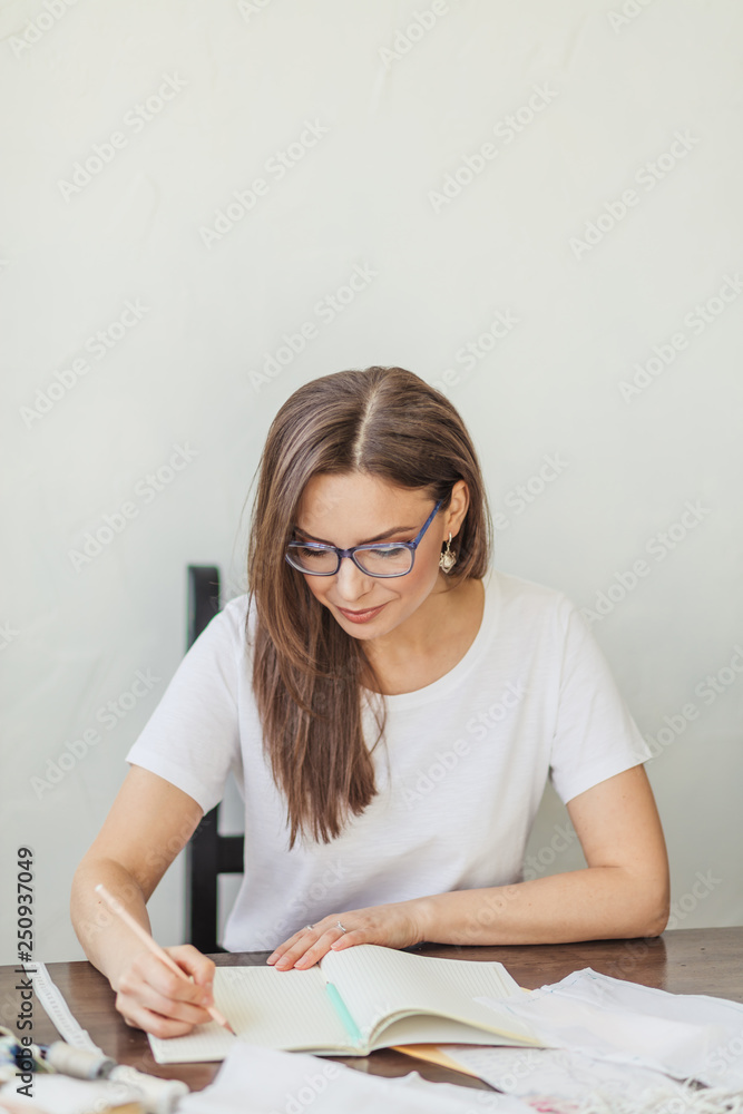 Skilled beautiful dressmaker in fashion atelier in stylish eyeglasses and white casual t-shirt drawing sketch of wedding dress for her female client Skilled beautiful dressmaker in fashion atelier in stylish eyeglasses and white casual t-shirt drawing sketch of wedding dress for her female client