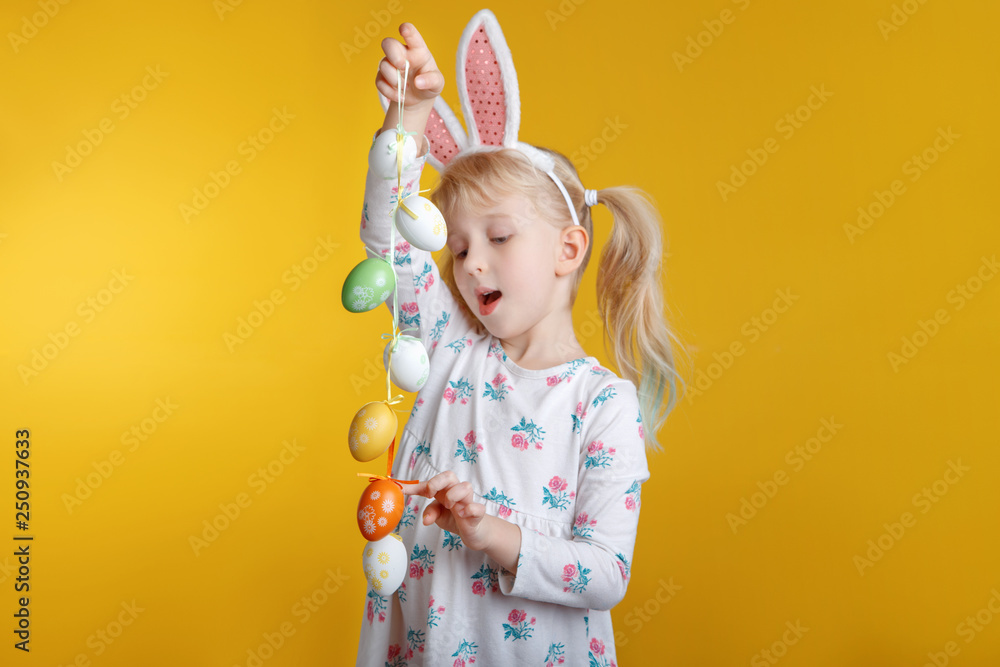 Cute adorable Caucasian blonde girl in white dress with pink  Easter bunny ears playing with eggs in studio on yellow background. Funny kid child celebrating traditional Christian holiday