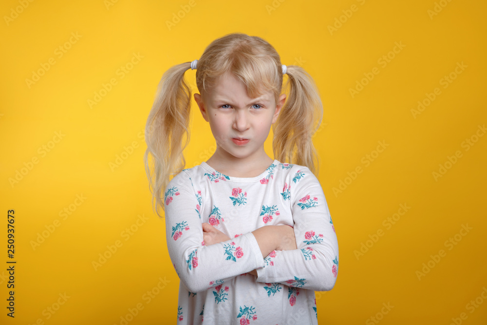 Cute grumpy angry Caucasian blonde girl with blue eyes in white dress posing in studio on yellow background with arms crossed on her chest. Kid child expressing negative emotion. Cute grumpy angry Caucasian blonde girl with blue eyes in white dress posing in studio on yellow background with arms crossed on her chest. Kid child expressing negative emotion.