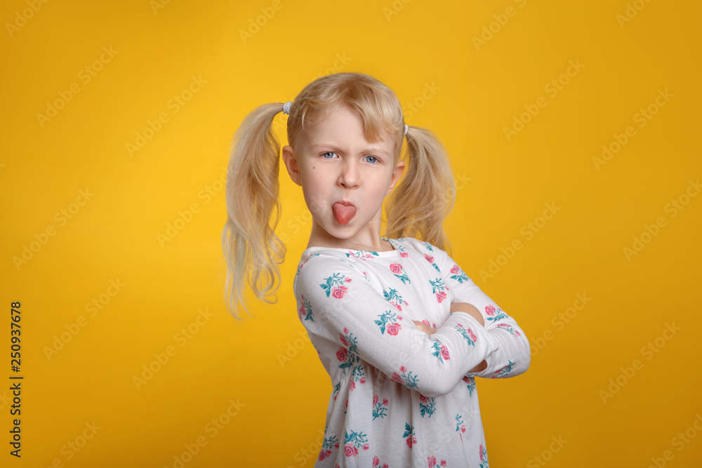 Grumpy angry Caucasian blonde girl with blue eyes in white dress posing in studio on yellow background with arms crossed on chest. Kid child expressing negative emotion showing tongue Grumpy angry Caucasian blonde girl with blue eyes in white dress posing in studio on yellow background with arms crossed on chest. Kid child expressing negative emotion showing tongue