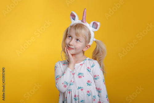 Cute adorable pensive Caucasian blonde girl in white dress wearing unicorn headband horn and ears thinking dreaming. Funny kid child expressing emotion standing in studio on yellow background
