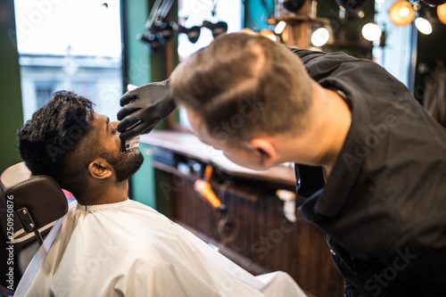 Grooming of real man. Young indian bearded man getting beard haircut at hairdresser while sitting in chair at barbershop