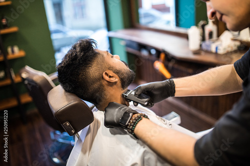 Barber shaving the beard of a handsome bearded man with an electric razor at the barber shop