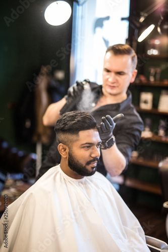 Barber combs his hair to a young guy with a beard and mustache and sprinkles hair on the spray gun. Barber Shop.