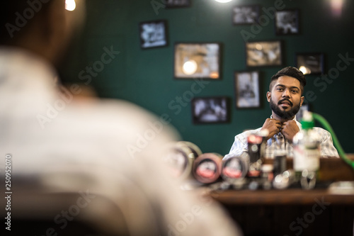 Time for new haircut. Handsome young bearded man looking at his reflection in the mirror and keeping hand in hair while sitting in chair at barbershop