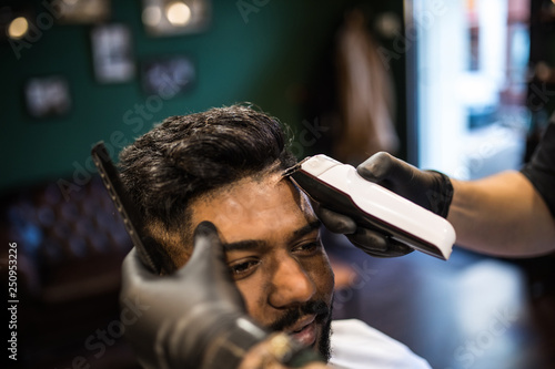 Perfect trim at barbershop. Young bearded man getting haircut by hairdresser with electric razor while sitting in chair at barbershop