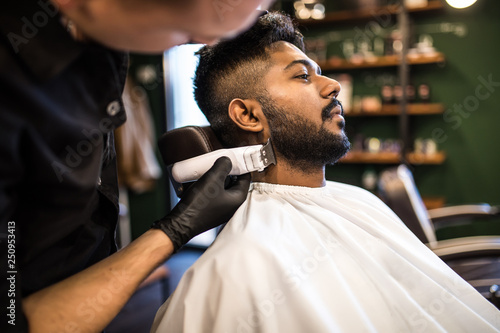 Perfect trim at barbershop. Young bearded man getting haircut by hairdresser with electric razor while sitting in chair at barbershop