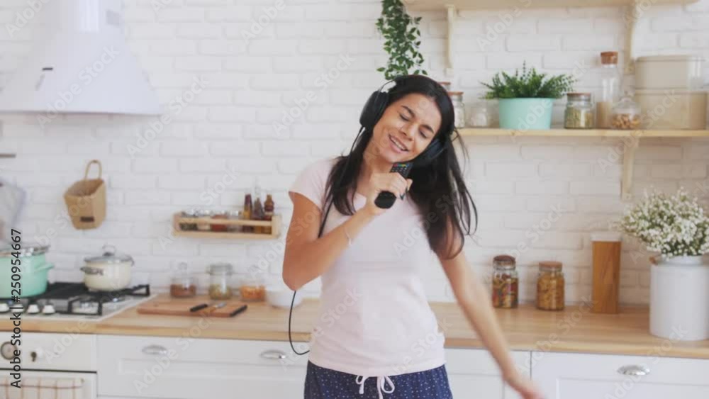 Young happy woman dancing and singing in kitchen in headphones and having some fun Young happy woman dancing and singing in kitchen in headphones and having some fun