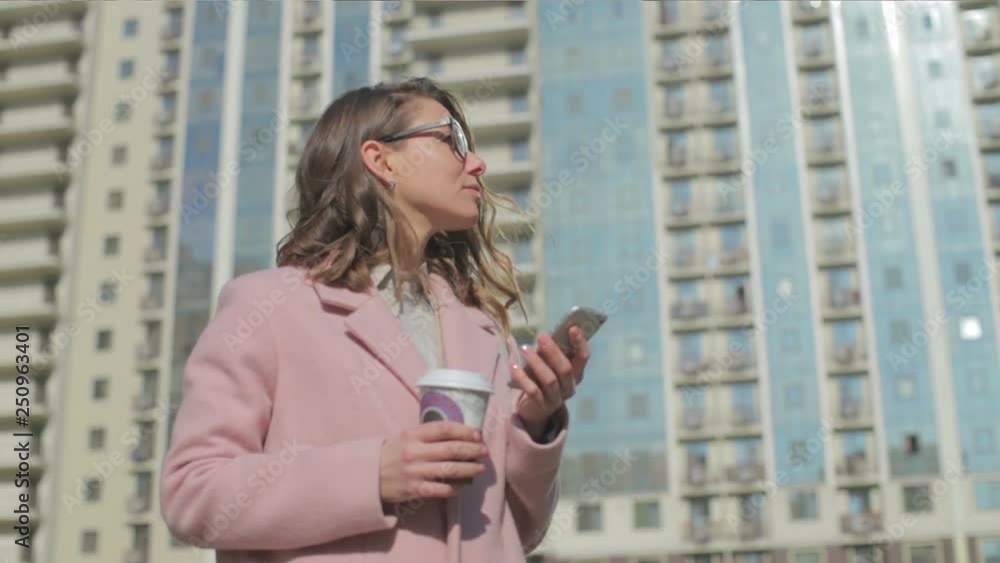 slow motion pretty blond lady in glasses wearing pink coat with phone coffee cup stands against building in sunny spring slow motion pretty blond lady in glasses wearing pink coat with phone coffee cup stands against building in sunny spring