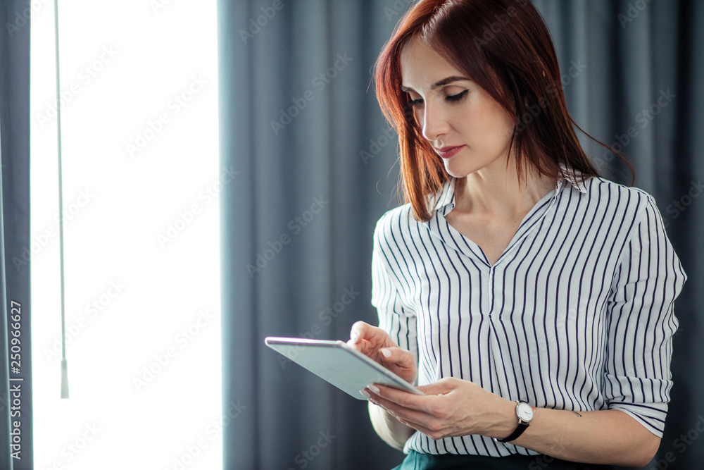 Fototapeta premium Red-haired business woman writing notes in her diary book while waiting her colleagues in office, sitting against window with grey curtains.