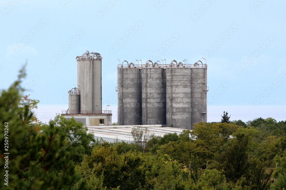 Tall concrete storage silos above industrial complex surrounded with dense forest vegetation and sea with clear blue sky in background on warm sunny day