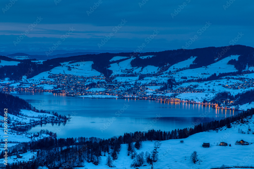 See mit beleuchtetem Dorf in der Dämmerung im Winter