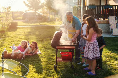 Grandfather teaching his grandchildren how to grill meal. Family gathering in backyard concept.