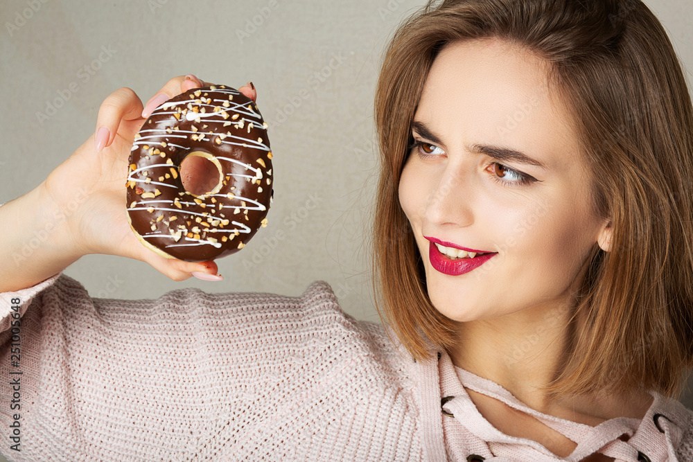 Young happy girl with pink lips holding chocolate donut over a grey background Young happy girl with pink lips holding chocolate donut over a grey background