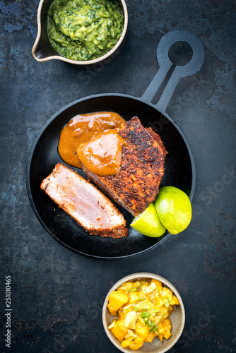 Traditional exotically blackened Cajun style tuna fish fillet with mango avocado salad, chimichurri dip and spicy BBQ sauce as top view in a modern design cast-iron pan