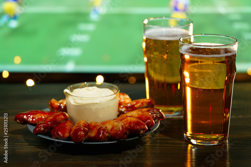 Hot barbecue chicken wings with 2 beer glasses on a dark wooden table served with honey mustard sauce. Football on a background, high resolution