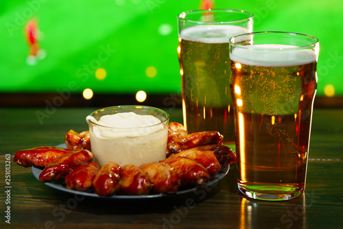 Hot barbecue chicken wings with 2 beer glasses on a dark wooden table served with honey mustard sauce. Football on a background, high resolution