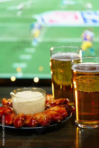 Hot barbecue chicken wings with 2 beer glasses on a dark wooden table served with honey mustard sauce. Football on a background, high resolution