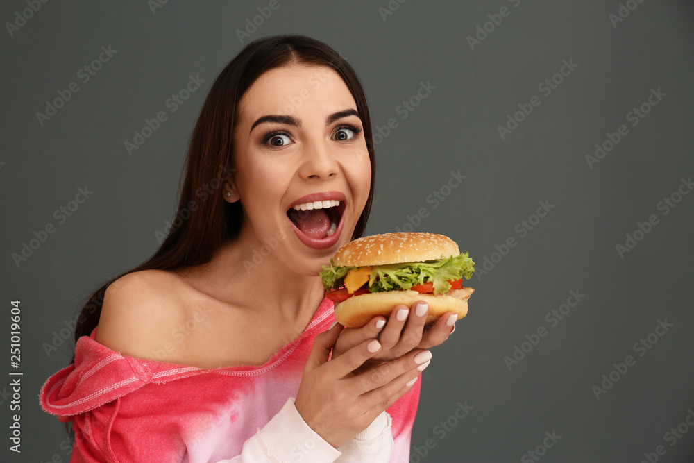 Young woman eating tasty burger on grey background. Space for text