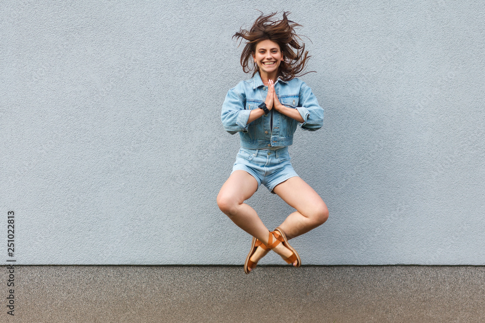 Free happy beautiful woman in casual jeans denim style in summertime jumping at light blue wall with namaste palm hands and looking at camera with toothy smile.