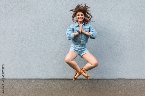 Free happy beautiful woman in casual jeans denim style in summertime jumping at light blue wall with namaste palm hands and looking at camera with toothy smile.