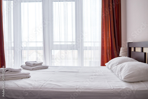 Hotel bedroom interior with empty double bed with wooden headboard and big window  copy space. White sheet  soft pillows and towels on bed  side view