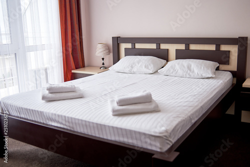 Hotel bedroom interior with empty double bed with wooden headboard, bedside table and big window, copy space. White sheet, soft pillows and towels on bed
