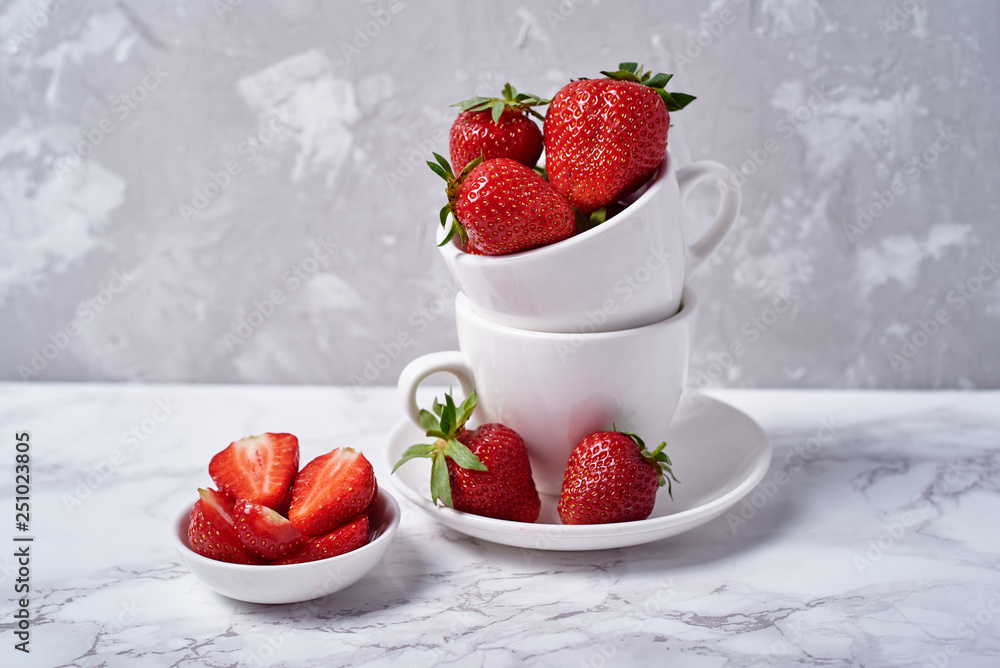 Red organic strawberries in white ceramic cup and strawberry slices in bowl on gray concrete background, copy space. Healthy food concept, still life Red organic strawberries in white ceramic cup and strawberry slices in bowl on gray concrete background, copy space. Healthy food concept, still life