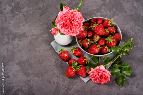 Red ripe strawberries in ceramic bowl on linen table napkin and pink rose in vase on gray concrete background, copy space. Healthy food concept, still life. Top view, flat lay