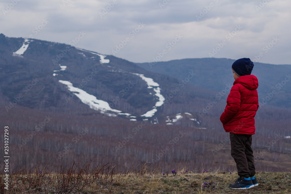 six-year-old child in warm clothes in full growth stands on the background of high mountains, foggy spring landscape
