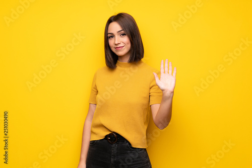 Young woman over yellow wall saluting with hand with happy expression © luismolinero