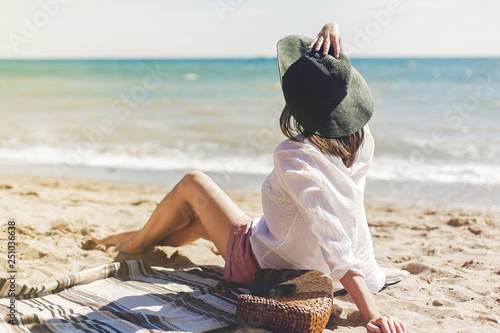 Summer vacation concept. Happy young woman relaxing on beach. Hipster slim girl in white shirt and hat sitting and tanning on beach near sea with waves, sunny warm weather. Peaceful calm moment