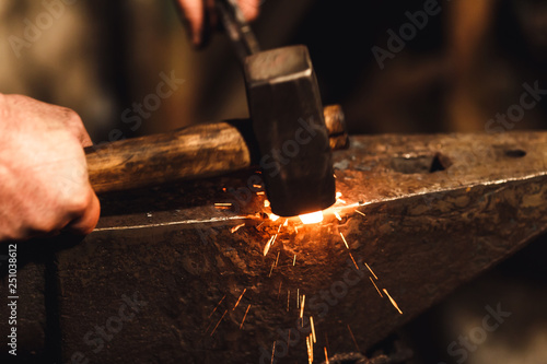 The blacksmith manually forging the red-hot metal on the anvil in smithy with spark fireworks.