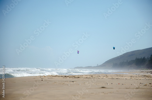 Sea beach with strong surf. Rocky cape, strong wind bends the trees and spreads a spray of waves. In the background are the kitesurfing sails in the sky. Sunny day.