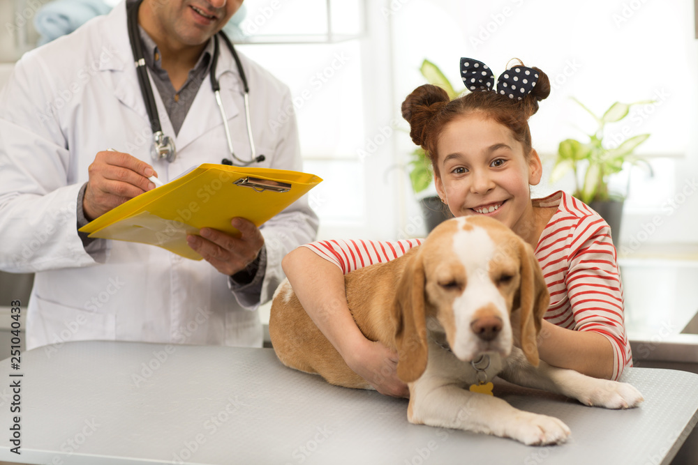 Horizontal shot of a cute happy little girl and her adorable beagle dog at the vet clinic copy space. Professional veterinarian making notes in his papers after medical examination on the back