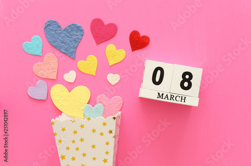 wooden March 8 calendar next to paper colorful hearts over pink wooden background