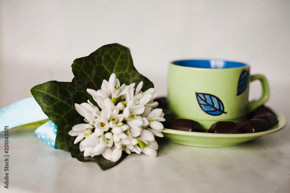 A cup of tea with pieces of black chocolate and a bunch of snowdrops on the table. A cup of tea with pieces of black chocolate and a bunch of snowdrops on the table.