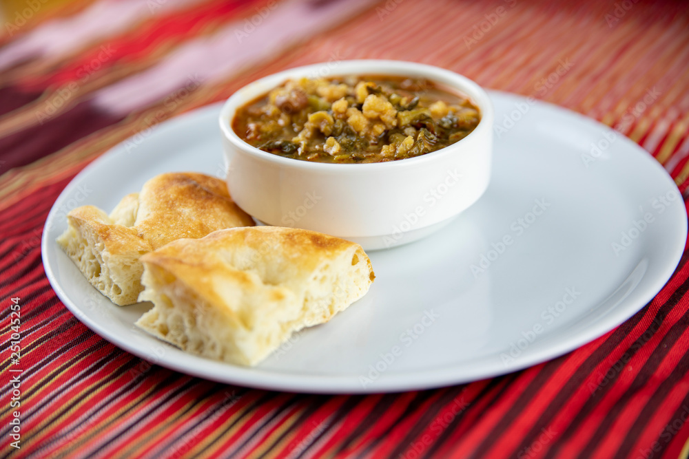 A bowl of hearty soup with chunks of vegetables and grains, served with pieces of crusty bread on a white plate, accompanied by a wooden utensil, set against a vibrant red and gold striped background. A bowl of hearty soup with chunks of vegetables and grains, served with pieces of crusty bread on a white plate, accompanied by a wooden utensil, set against a vibrant red and gold striped background.