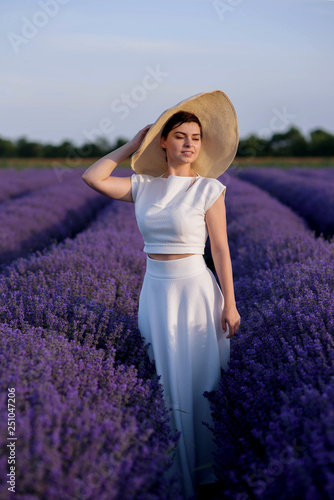 Elegant young woman in a white dress and a hat walking through a lavender field