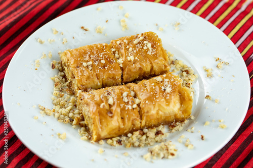 Two pieces of golden, flaky baklava on a white plate, garnished with crushed nuts, set against a vibrant red and gold striped background. photo