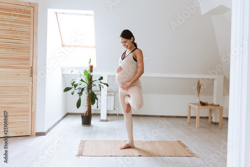 Young pregnant yoga woman working out at home doing prenatal Tree posture, Vrksasana yoga pose. Healthy Pregnancy concept. Pregnant Namaste gesture photo