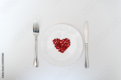 On a white plate from pomegranate seeds composite heart shape. Knife and fork next to the plate. White background. Flat layer.