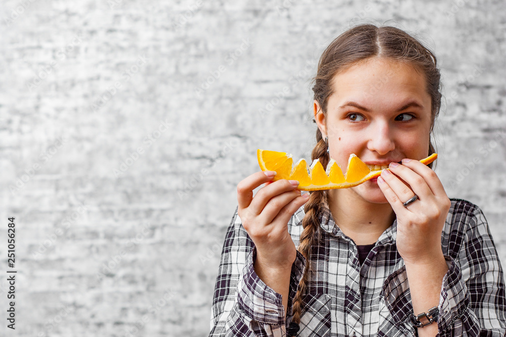 portrait of young teenager brunette girl with long hair eat orange fruit on gray wall background with copy space. portrait of young teenager brunette girl with long hair eat orange fruit on gray wall background with copy space.
