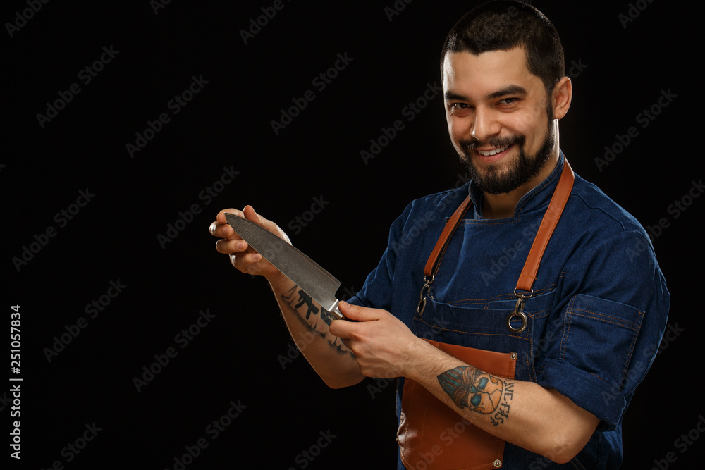 Positivity and smiling chef in brown apron demonstrating metallic knife, showing and looking at camera. Professional male working with cooking equipment. Isolate on black background. Positivity and smiling chef in brown apron demonstrating metallic knife, showing and looking at camera. Professional male working with cooking equipment. Isolate on black background.