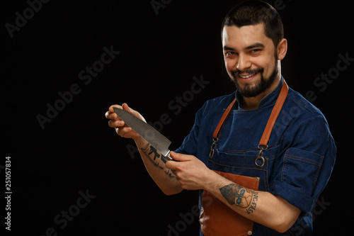 Positivity and smiling chef in brown apron demonstrating metallic knife, showing and looking at camera. Professional male working with cooking equipment. Isolate on black background. © Nestor