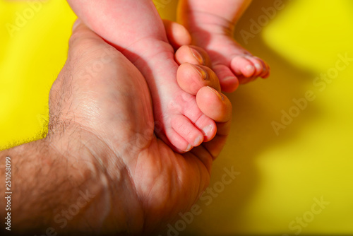 Baby legs on a yellow background, dad holds the small legs of a newborn baby in his hand.