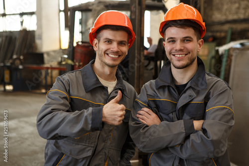 Quadro em tela Portrait of two happy workers of metal stock in orange helmets and gray uniform standing together, posing and looking at camera