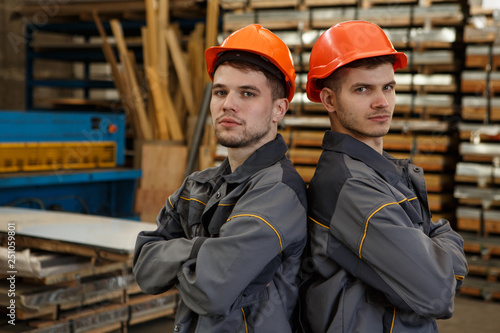 Portrait of two workers wearing orange helmets and gray protective suits standing back to back. Serious colleagues standing with arms folded, posing and looking at camera. Concept of job and teamwork.