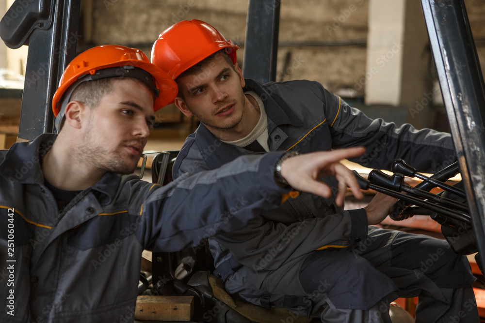 Side view of two loaders in orange helmets and gray suits talking on metal stock. One worker pointing with finger at materials and another young man sitting on forklift, listening and looking there. Side view of two loaders in orange helmets and gray suits talking on metal stock. One worker pointing with finger at materials and another young man sitting on forklift, listening and looking there.